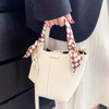 A cream handbag with a checked silk chiffon scarf tied to the handle, held by a hand with red nails.