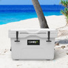 A white 25L cooler box with a logo, set on a sandy beach near palm trees and blue water.