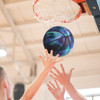 A basketball in shades of blue and purple is flying towards a hoop, within a gymnasium setting. The scene shows hands reaching up to catch it.