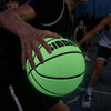 A glowing green basketball with a textured surface, held in a player's hand. The background features blurred players.