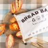 A reusable linen bread bag in natural fabric, surrounded by various loaves of bread on a woven mat.