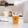 A glass tumbler filled with beer, showcasing a golden amber colour and bubbles, with a branding logo visible.