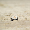 A white and black Hacky Sack is resting on a sandy surface, featuring a logo on its front.