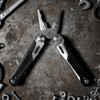 A black and silver multi-tool with pliers and various tools, surrounded by metal tools and screws. It features a logo.