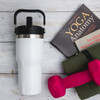 A drink bottle with a white body and black lid, alongside a pink dumbbell and yoga book on a wooden surface.