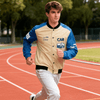 A young man runs on a track wearing a beige and blue varsity jacket with a logo on the chest.