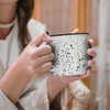 A ceramic mug featuring a speckled black and white design, held by a person with one hand. The mug has a contrasting rim.