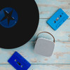 A white mini speaker with a brown handle, accompanied by blue cassette tapes and a black vinyl record on a wooden surface.