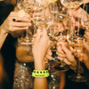A vibrant, yellow PVC wristband with a logo appears among hands holding champagne glasses at a celebratory event.