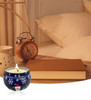 A blue tin candle with a holiday design, surrounded by a bedside table with a clock and a book.