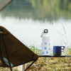 A drink bottle in white with a logo, placed on a table near a camping chair by the water.