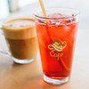 A glass filled with a bright red drink and ice, alongside a cup of frothy coffee. The glass features a logo.