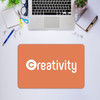 An orange rubber counter mat with the word "creativity" printed on it, surrounded by office supplies and a laptop.