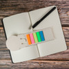A ruler-shaped bookmark in cream with five vibrant sticky notes in pink, orange, green, and blue, on an open notebook with a pen.