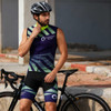 Men's sublimated cycling shorts in blue and green with a fitted design. The model wears a helmet and leans against a bike.