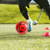 A vibrant red soccer ball with black and orange designs on grass, with a person in action nearby.