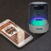 A portable Bluetooth speaker with a blue and green light display next to a smartphone displaying a music app.