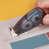 A hand using a transparent, grey Metz Glue Tape dispenser on paper, displaying a smooth adhesive application.