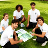 A group of five young adults wearing unisex white RPET sublimated round-collar tee shirts, sitting on grass.