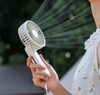 A white foldable USB hand fan is held in a person's hand, with air blowing from its grille. It features a logo.