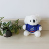 A white koala plush toy wearing a blue shirt, sitting beside a small potted plant on a wooden surface.