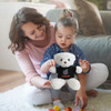A white plush bear wearing a black shirt sits on a floor with a woman and a child playing together.
