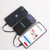 A black power bank with a logo, charging two smartphones on a light surface. The power bank has a sleek design.
