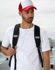 A black backpack worn by a man in a white t-shirt and a red and white cap, with earbuds visible.