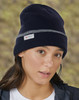 A navy beanie with a grey band and a logo, worn by a person with long hair in a blurred outdoor background.