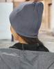 A grey cable knit beanie, viewed from the back, worn by a person in a jacket outdoors.