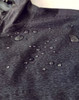 A close-up of a dark grey waterproof jacket with water droplets on the fabric and a hood. The jacket has a logo.