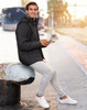 A man wearing a black unisex sustainable parka jacket, seated by a waterfront, holding a phone.