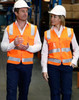 Two individuals wear bright orange Hi-Vis safety vests with reflective tapes and hard hats in a warehouse setting.