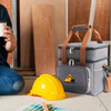 A grey 10L cooler bag with a logo, placed beside a yellow hard hat and a person holding a drink.