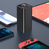 A black power bank stands upright, surrounded by smartphones and a gaming console on a dark surface.