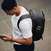 A black backpack is worn by a person looking at their phone, with a drink bottle visible in a side pocket.