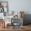 A foldable metal laptop stand in grey, displayed on a wooden desk alongside a laptop and stationery.