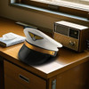 A pilot-themed cap in white and black, adorned with gold detailing, resting on a wooden desk next to a vintage radio.