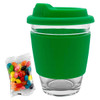 A glass coffee cup with a green silicone lid and sleeve, accompanied by a pack of colourful jelly beans.