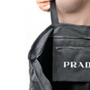 A black tote bag with an interior pocket and a visible logo. The bag's straps are being held by a hand.