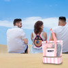 A pink and clear mesh beach tote bag with handles, placed on sand with three people sitting in the background.