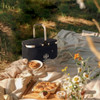 A black picnic cooler basket sits on a blanket, surrounded by flowers and food items. It has a silver handle and logo.