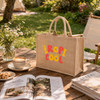 A natural jute tote bag in beige with a colourful logo, placed on a wooden table alongside coffee and magazines.