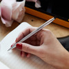 A silver ballpoint pen held in a hand, with a notebook and flowers in the background.