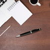 A black metal pen with a sleek design sits on a wooden desk alongside a notepad, pencil, laptop, and coffee cup.