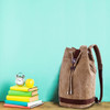 A brown canvas bucket backpack sits next to stacked books, an alarm clock, and a green apple against a turquoise background.