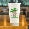 A white compostable paper cup with a logo featuring a cartoon character, placed on a wooden surface.