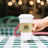 A white paper cup with a sleeve, featuring a food truck design, held by a hand against a checkered tablecloth backdrop.