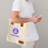 A cream-coloured canvas bag with brown accents and a logo, held by a person checking their phone.