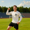 A person in a white long-sleeve crew neck shirt poses energetically on a sports field, wearing black shorts.